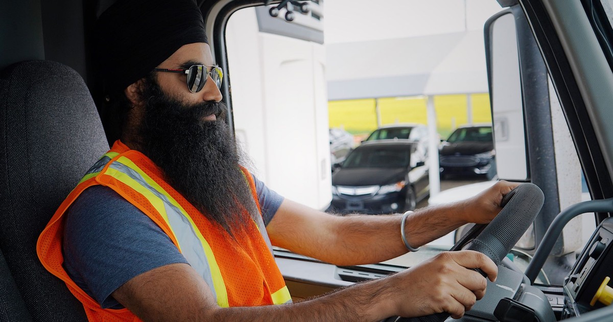 Bearded truck driver wearing a turban and safety vest inside a truck cab, focused on driving.