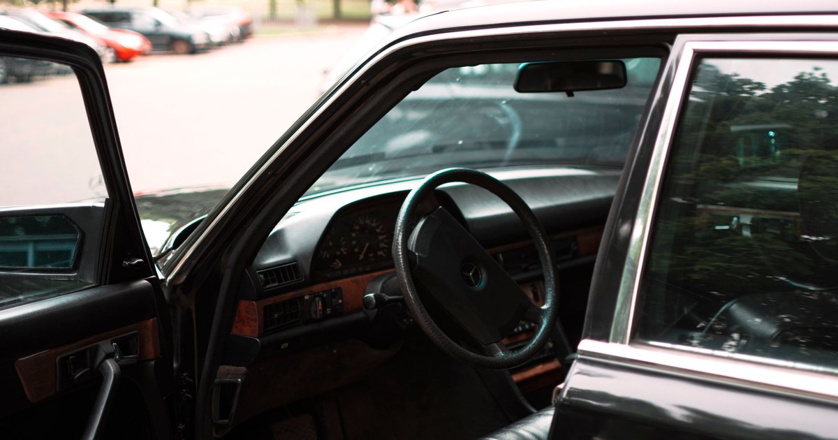 Close-up of an open car door showing the steering wheel and dashboard with a blurred parking lot background.