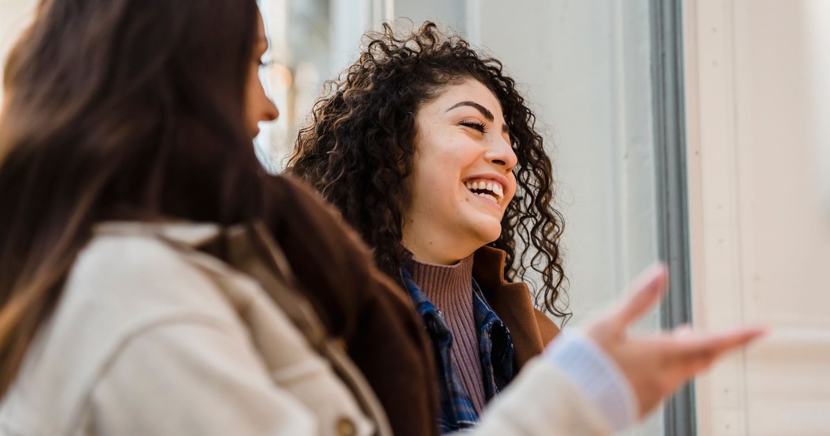 Side view of happy young multiethnic female best friends with long dark hairs in casual clothes laughing while looking at showcase of fashion store on city street