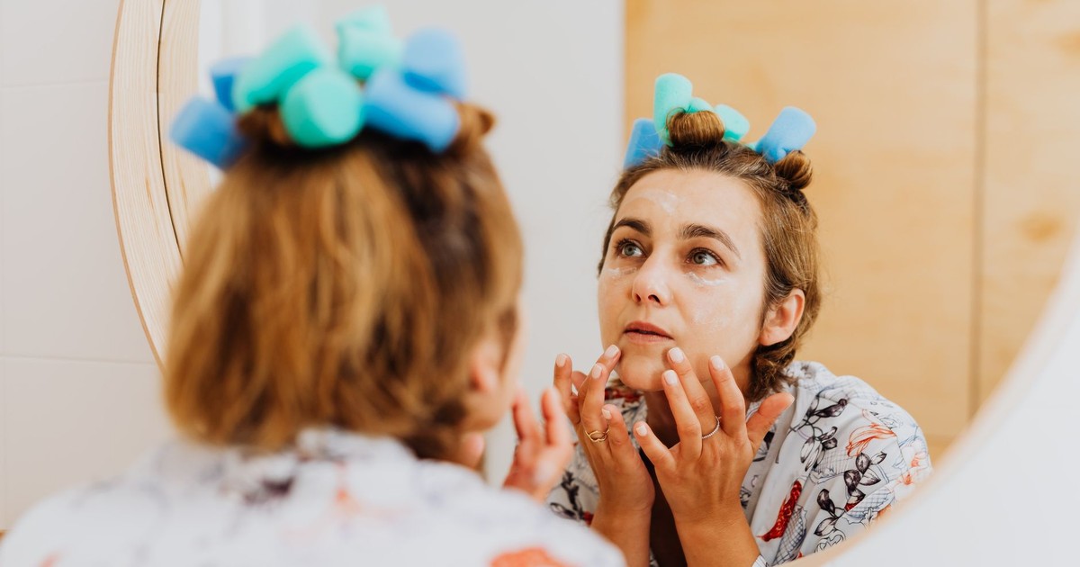 Woman with curlers applying cream in front of a mirror indoors.