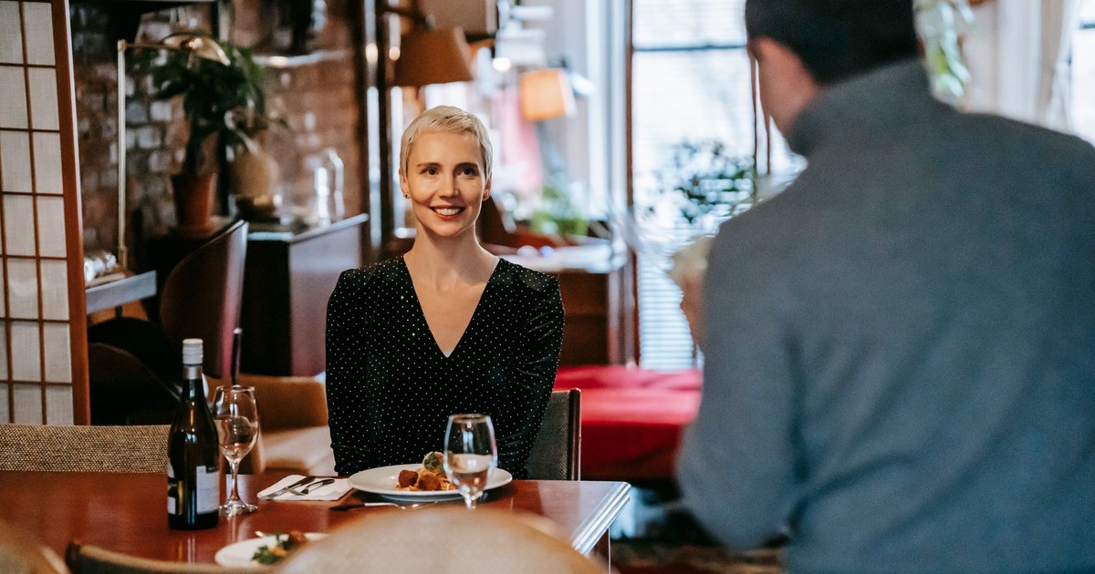 Positive adult couple having date in bright room while man standing with bouquet with flowers near chairs and table with glasses and wine bottle near plate with food and cutlery