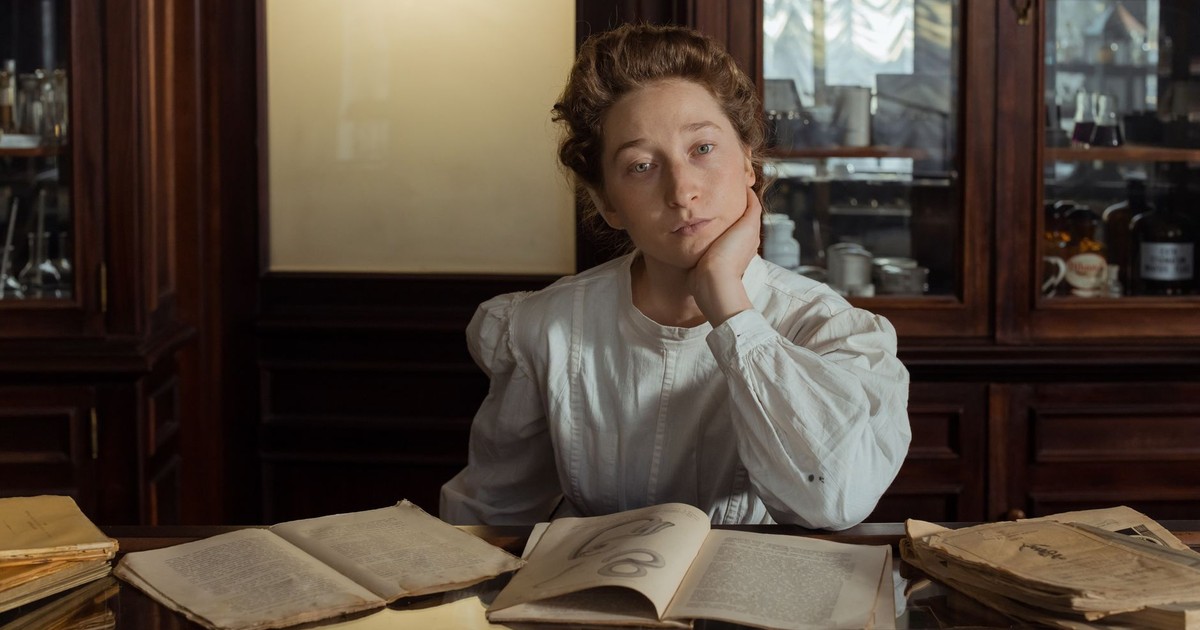 Woman in vintage attire reading old books at a wooden table in an antique-style room.