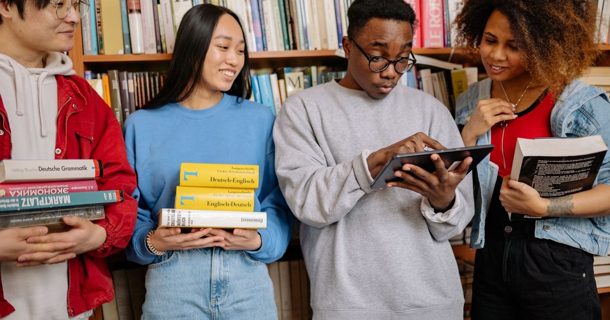 A group of diverse students study together in a library, using books and a tablet for learning.