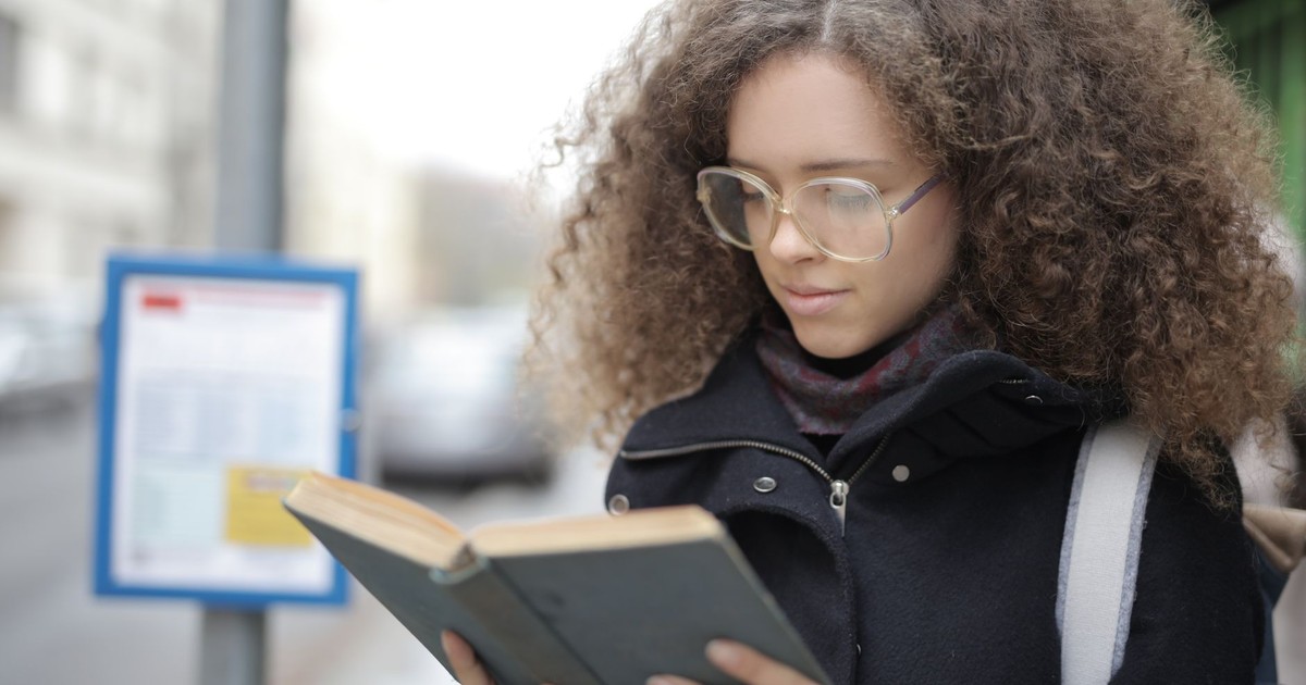 Serious young woman with curly hairs wearing warm clothes and eyeglasses reading book while waiting for bus on sidewalk