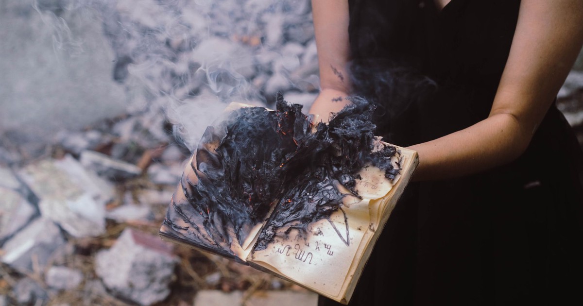 A woman holds a burning book, with smoke and ashes rising outdoors.
