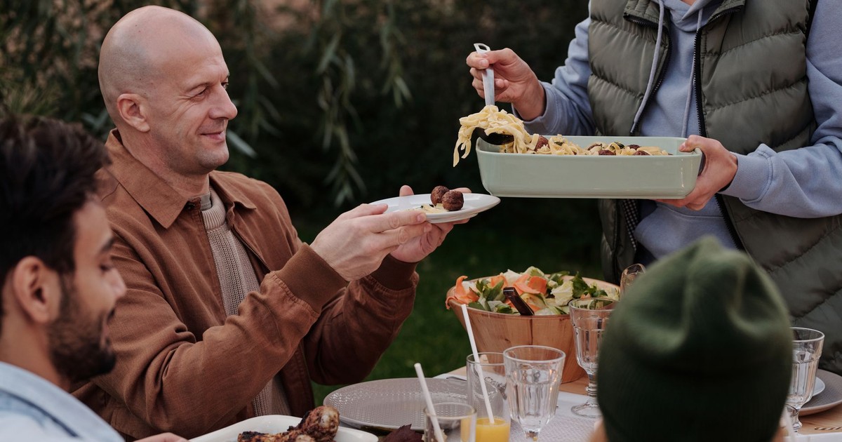 Faceless female in warm vest put pasta with meatballs on plate of bald man during dinner with friends outdoors