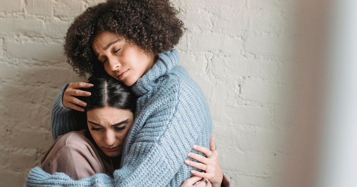 Sad African American woman with closed eyes hugging and consoling depressed female friend near white wall in light room at home
