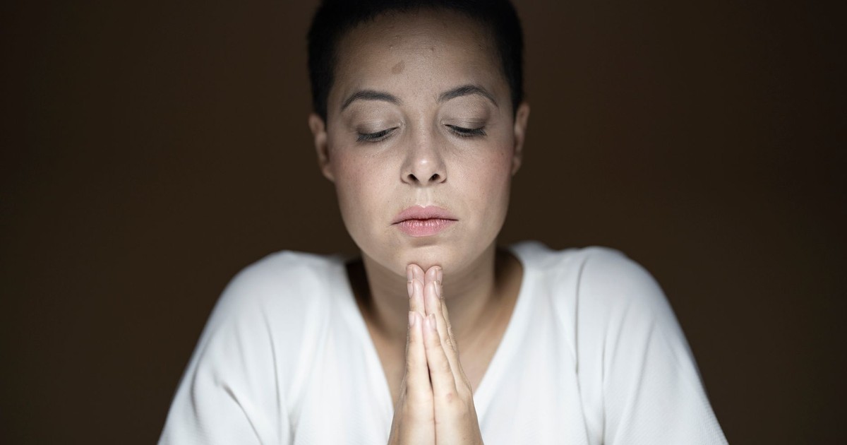 A peaceful woman with closed eyes and clasped hands in deep prayer inside a quiet room.