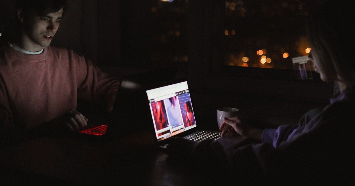 A man and woman using laptops at night, illuminated by screens in a dark, cozy room.