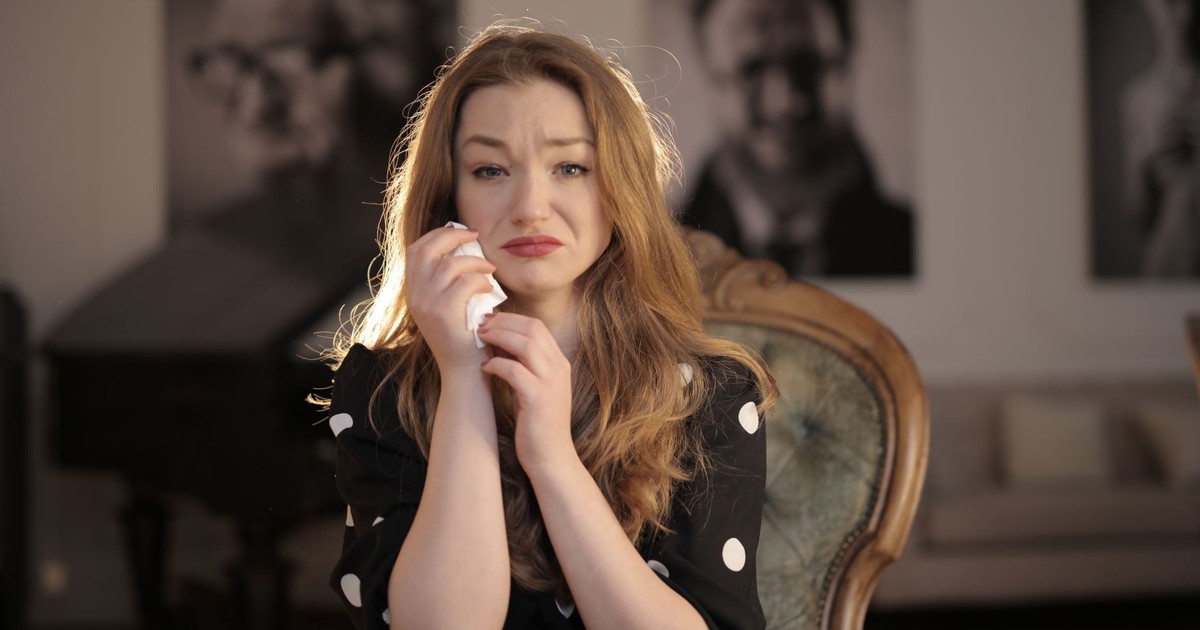 A woman sitting in a vintage chair indoors, showing emotions and holding tissues.