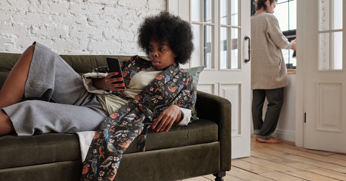 African American woman lounging on a sofa, texting, while man stands by window.