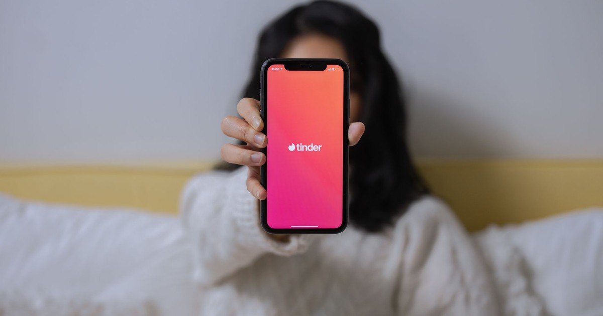 A woman in casual attire displays the Tinder app on her smartphone while sitting indoors.