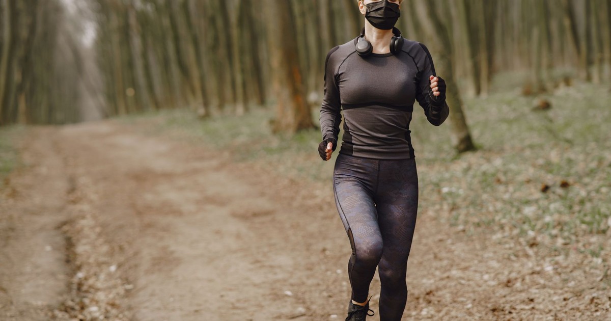 Full body sportswoman in activewear and medical mask with headphones on neck running through autumn forest with closed eyes during workout