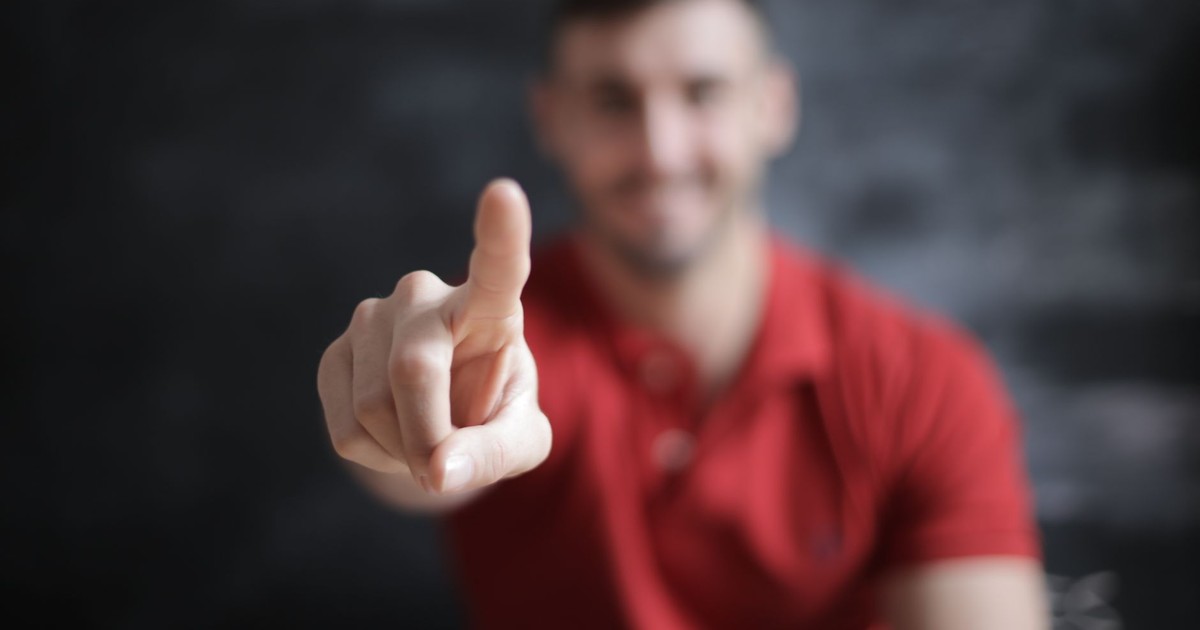 A man in a red shirt with a blurred face points his finger directly at the camera lens.