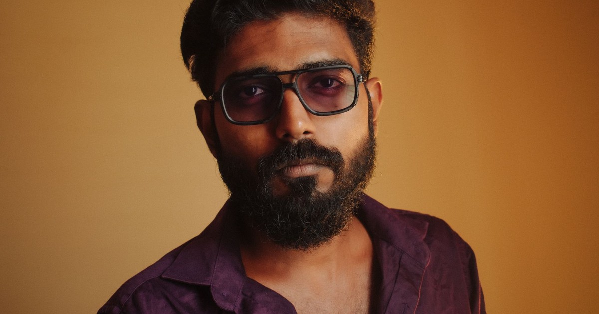 A close-up portrait of an adult man with a beard wearing sunglasses and a purple shirt.