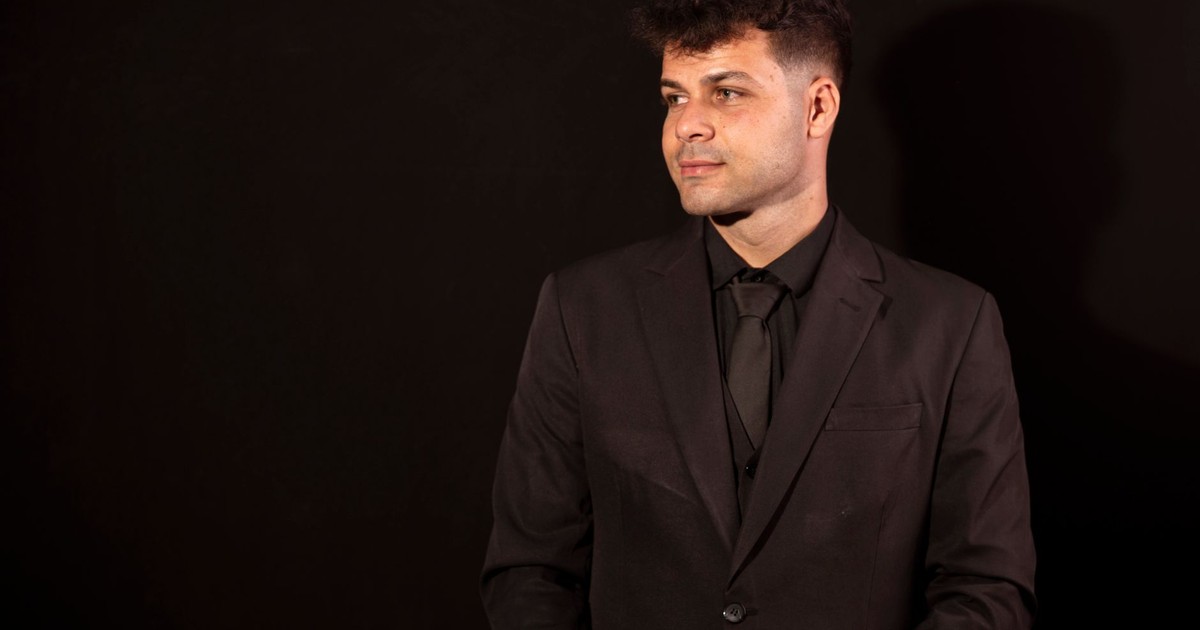 A confident man in a black suit poses against a dark background, exuding professionalism.