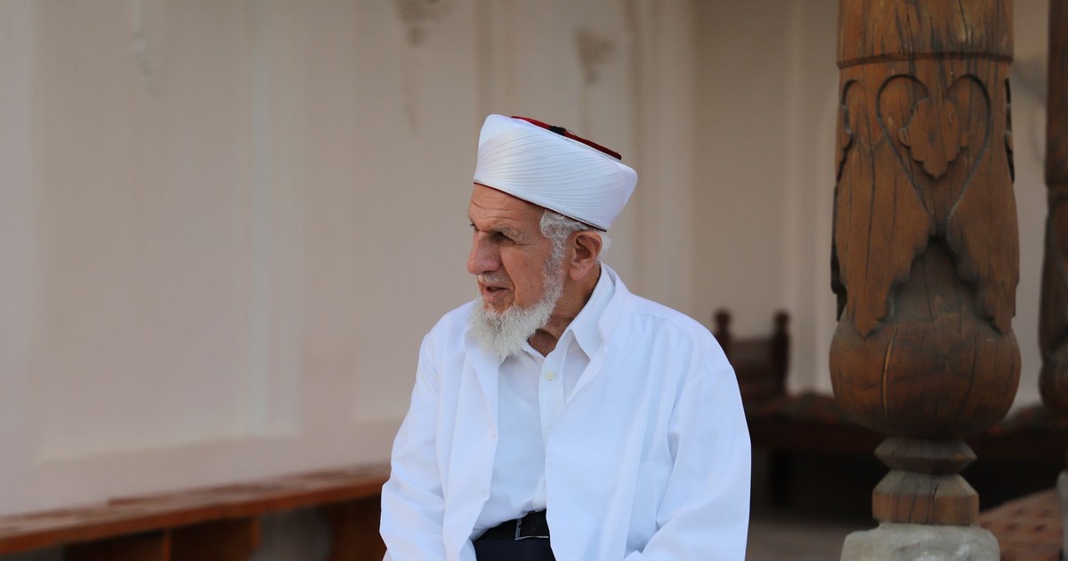 Senior man with white beard and hat in traditional attire sitting outdoors.