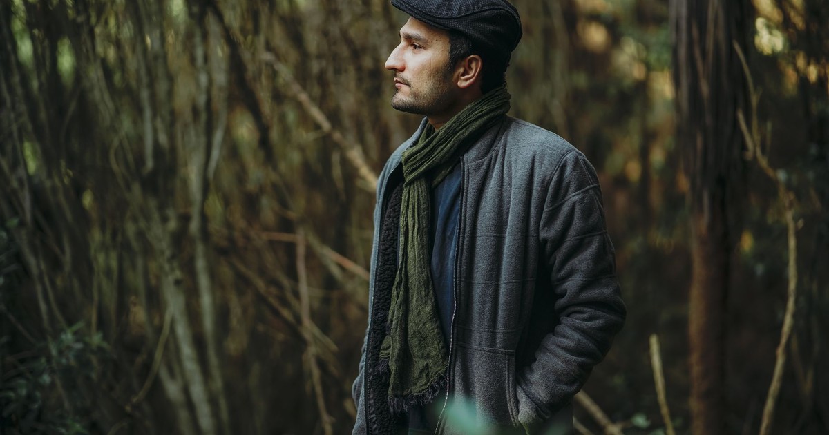 A man wearing a gray jacket and scarf poses in a lush forest, creating a contemplative mood.