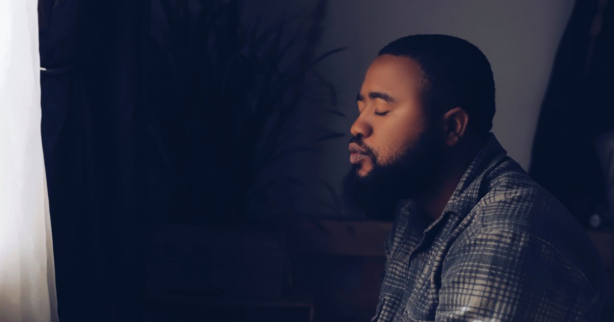 A man in a checkered jacket sits peacefully with eyes closed in a dimly lit room, evoking a sense of calm.