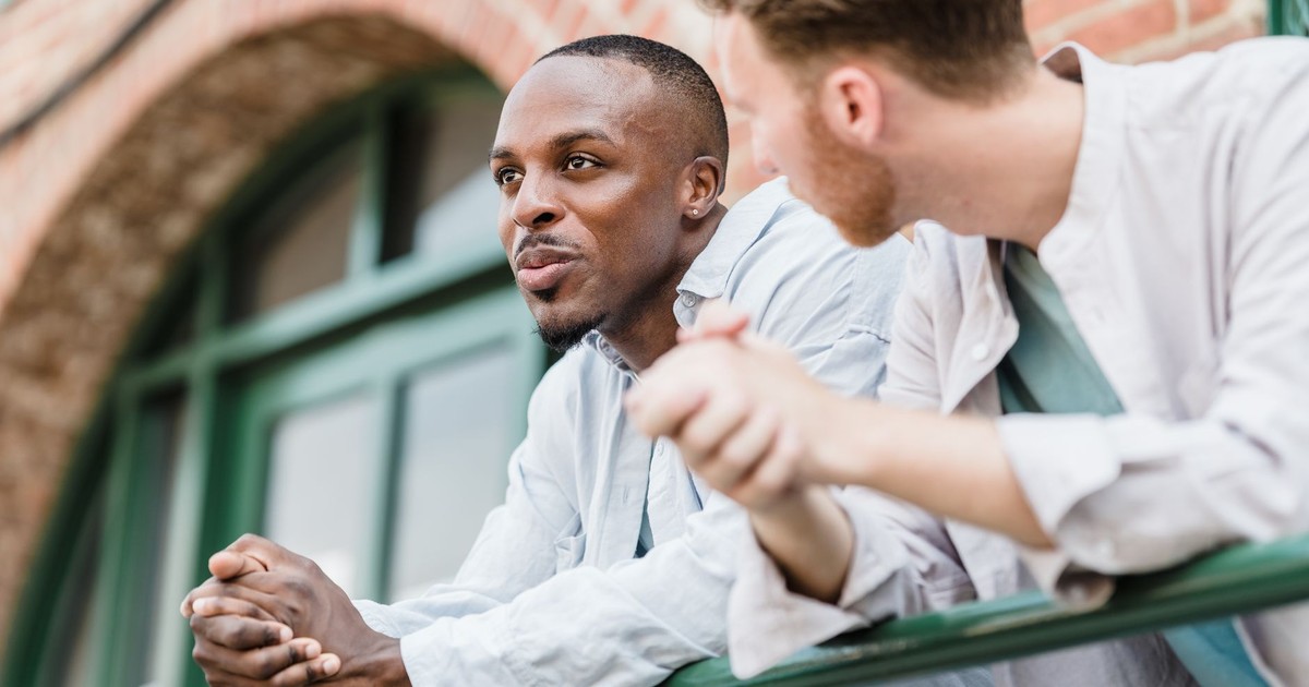 Two men lean on a railing outdoors, engaged in a relaxed conversation.