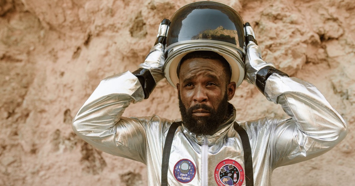 African American astronaut putting on helmet in rocky setting, wearing a shiny silver space suit.