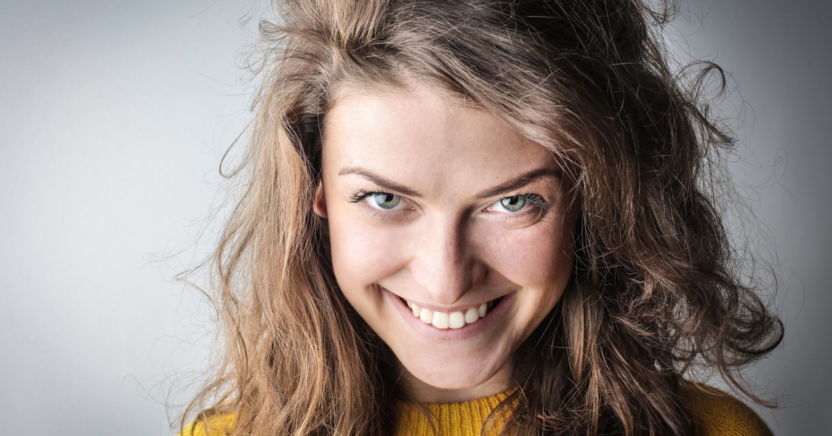 Close-up portrait of a smiling woman in a yellow sweater and casual hair, exuding happiness against a gray background.