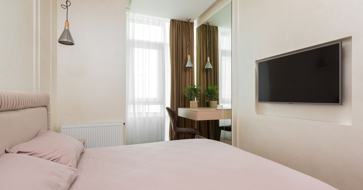 Comfortable compact bedroom with big mirror near small window with curtains and TV placed on white wall against soft bed in modern apartment