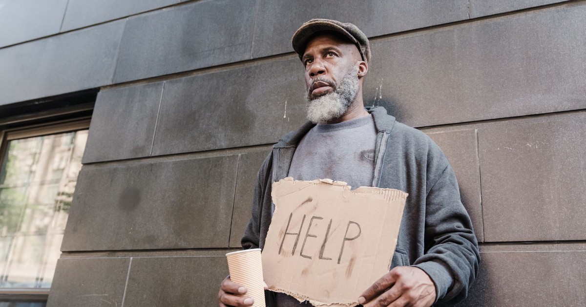 An elderly homeless man holds a cardboard sign asking for help in an urban setting.