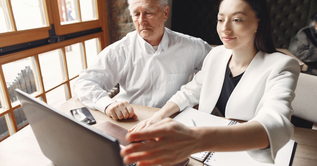 Modern adult woman with senior male executive browsing laptop and checking notes while working remotely in creative workspace near window and looking at screen