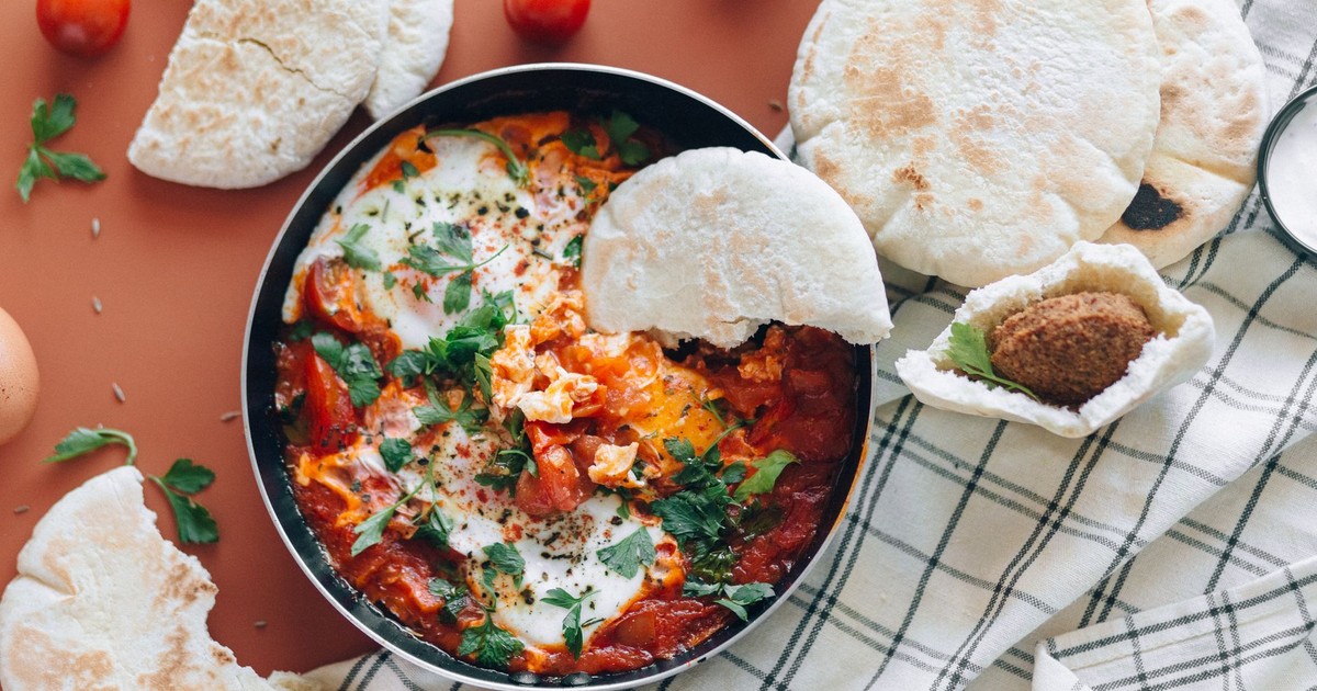 Top view of shakshuka meal with pita bread, cherry tomatoes, and fresh herbs.