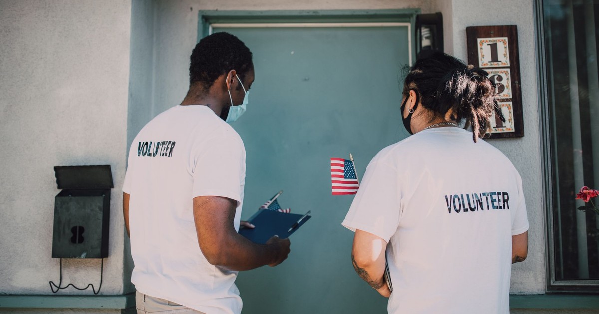 Two volunteers wear masks and visit community homes, engaging in outreach activities.