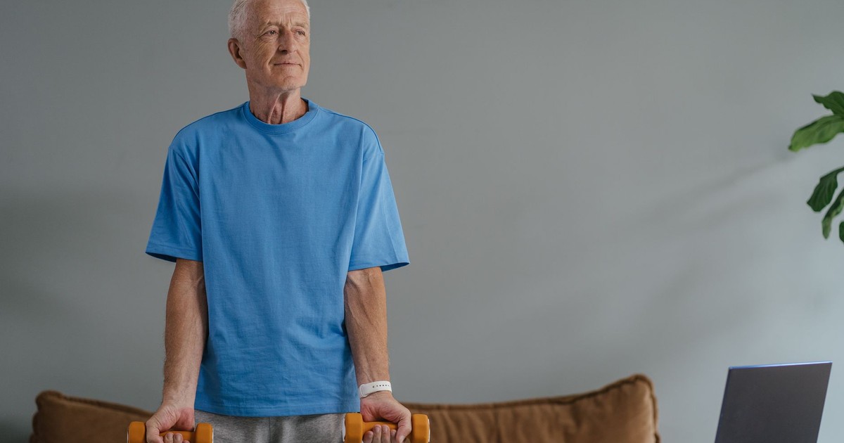 Elderly man in blue shirt lifting dumbbells near laptop, embracing home fitness.