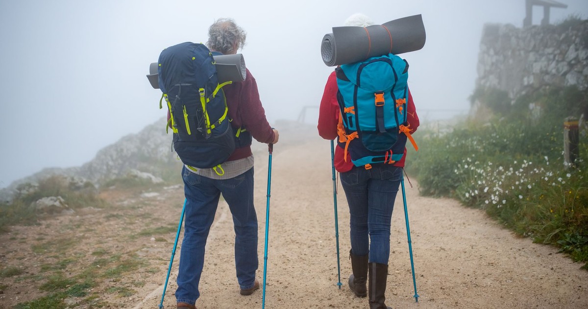 Elderly couple hiking on a foggy trail in Portugal, equipped with backpacks and walking poles.