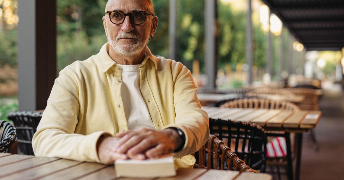 Elderly man with eyeglasses reading at an outdoor cafe table, enjoying a peaceful setting.