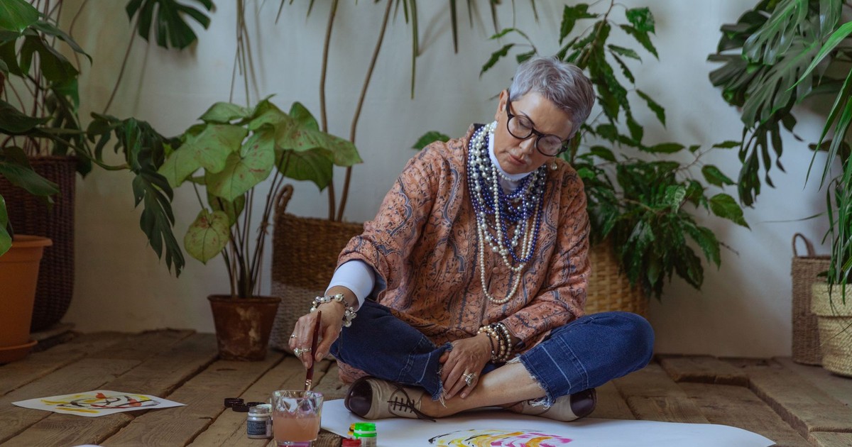 Elderly woman painting watercolors surrounded by plants indoors, artistically focused.