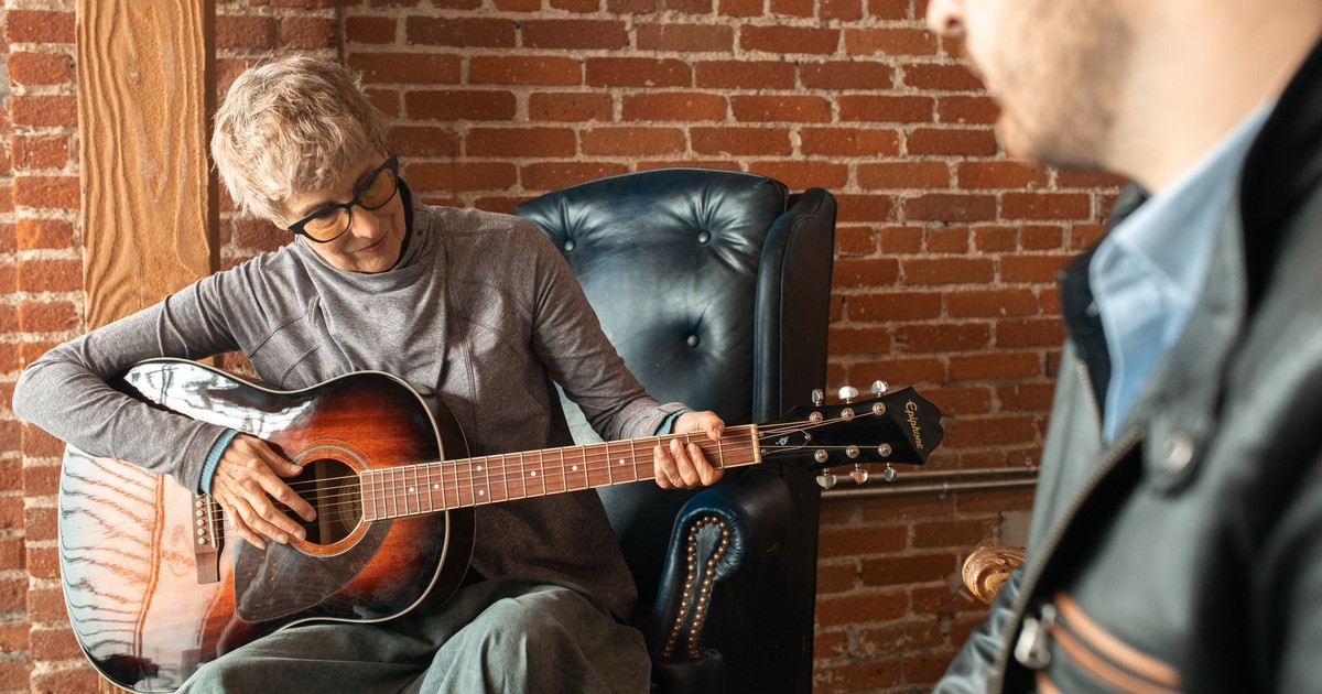 Senior woman enjoying playing acoustic guitar indoors with a companion nearby.