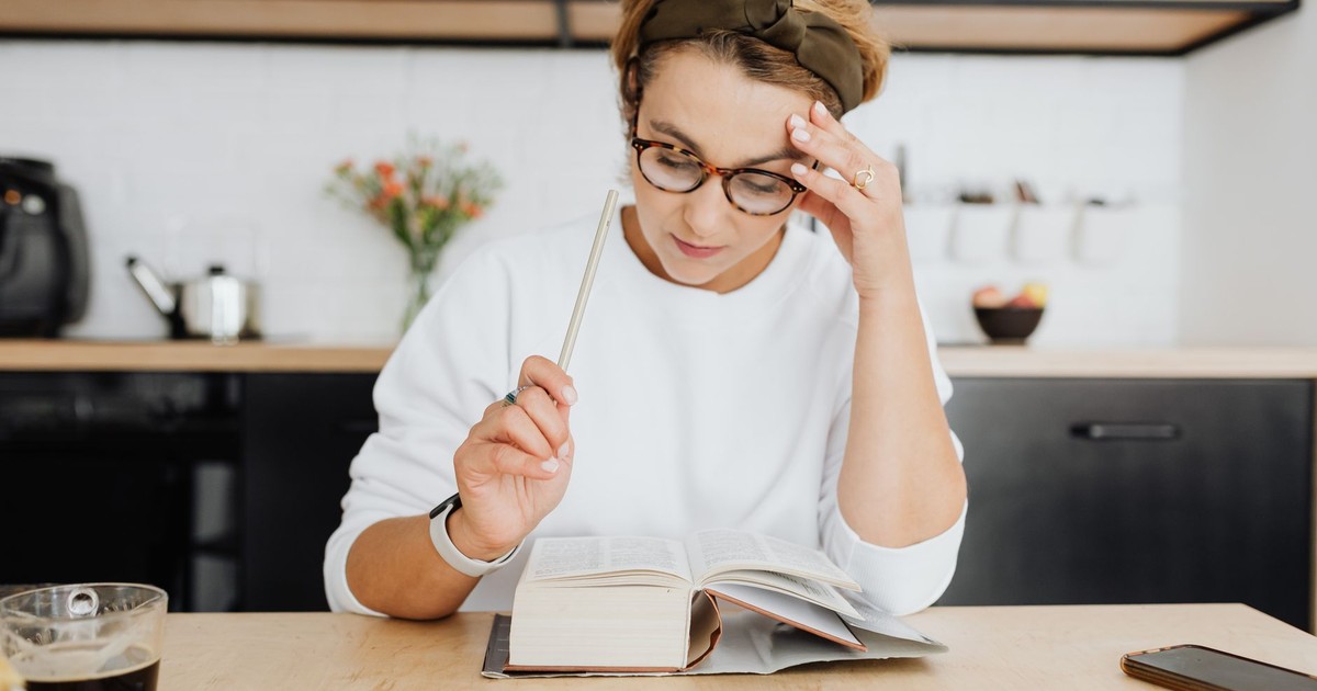 A focused woman in glasses reading a book in a bright, modern kitchen setting with coffee.