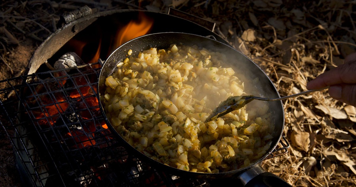 Rustic hash browns cooking on a campfire grill outdoors, emitting steam and aroma.