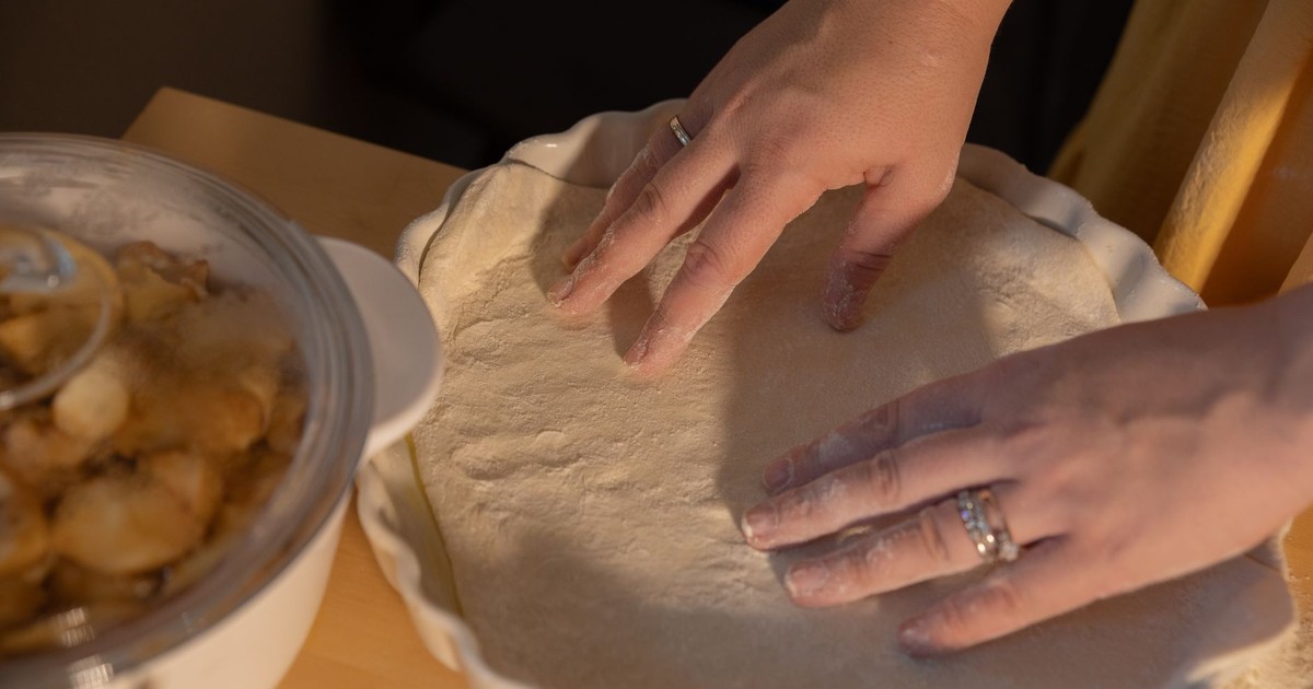Hands shaping dough in a pie dish next to apple filling, ready to bake.