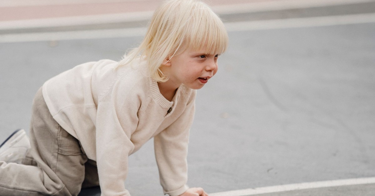 Side view of adorable cheerful little kid with blond hair standing on all fours on sports ground against blurred background