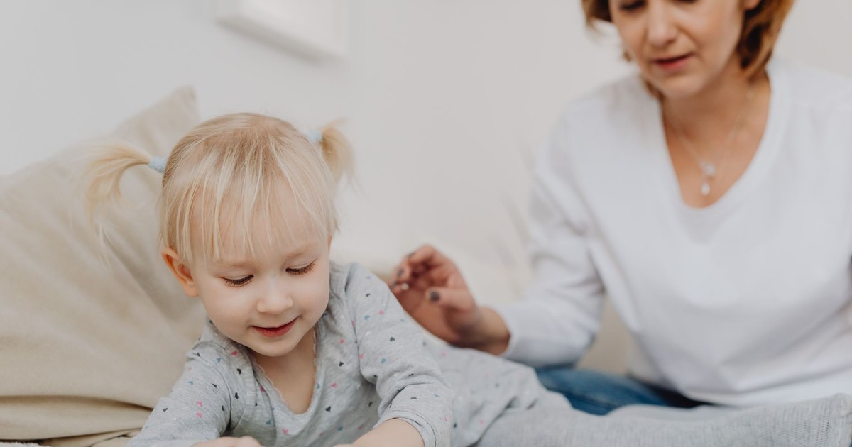 A smiling toddler with pigtails playing indoors with her mother watching.