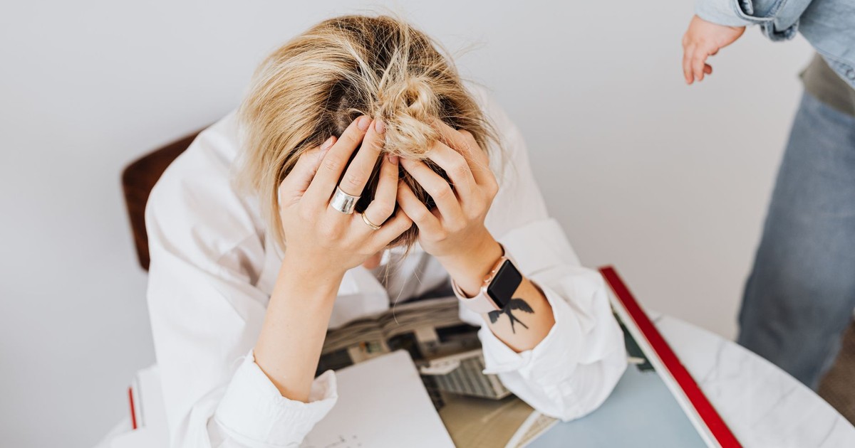 Frustrated woman in white shirt sitting with head in hands, overwhelmed by work tasks.