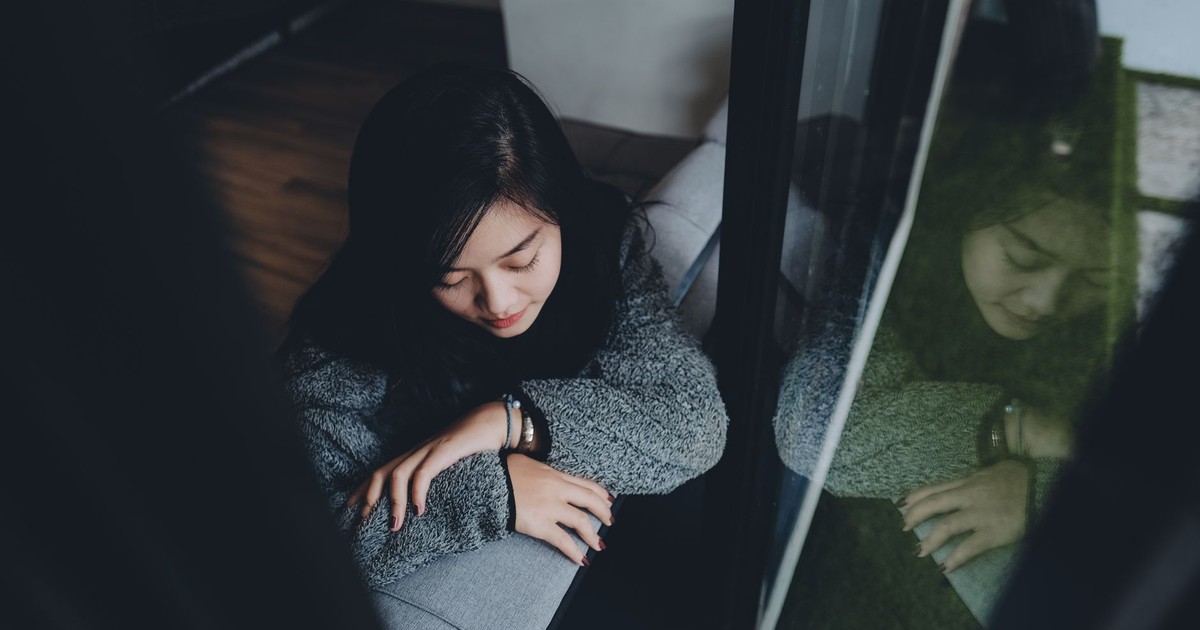 A young Indonesian woman in deep thought as she leans on a window sill indoors.