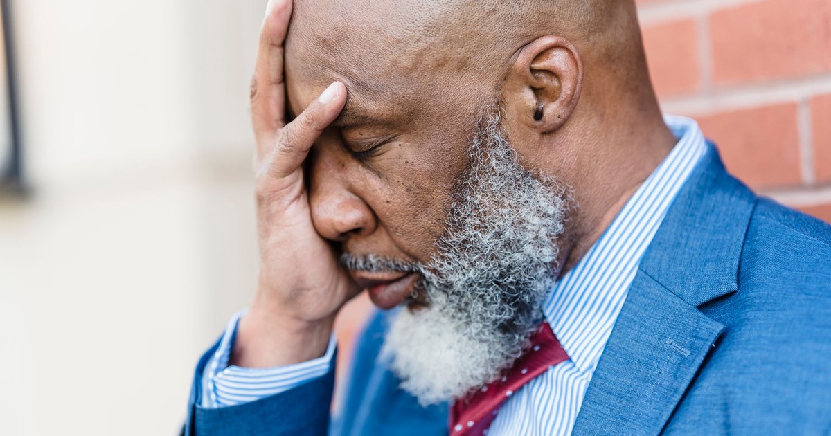 Side view of sad African American male entrepreneur in elegant suit covering face with hand while standing near building on street
