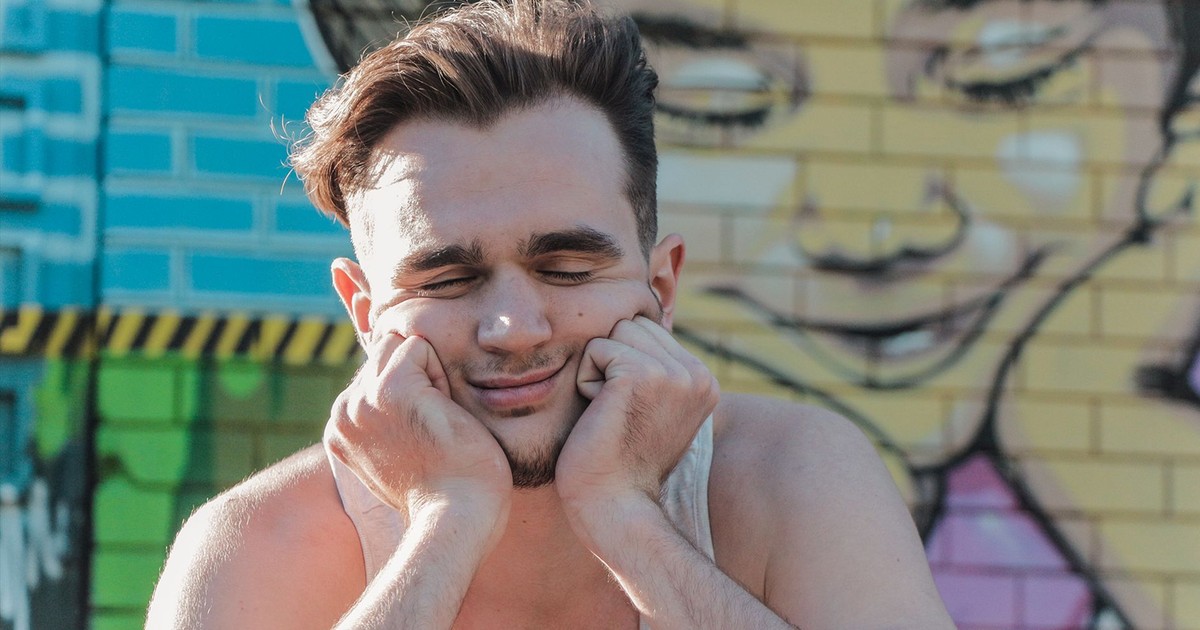 A man with closed eyes smiles playfully against a vibrant wall mural outdoors.