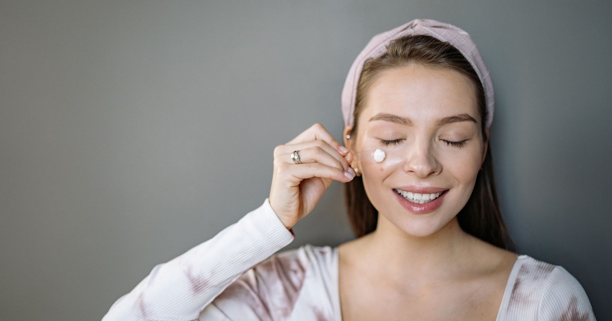 Smiling woman applying skincare cream with eyes closed on gray background.