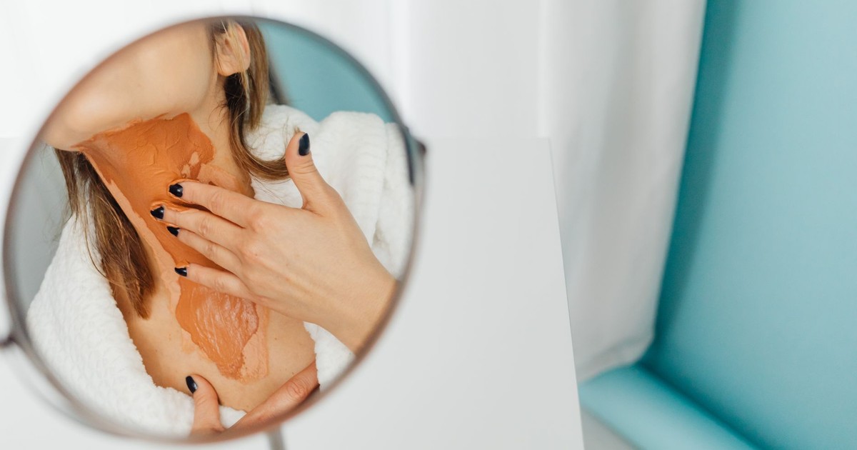 Close-up of a woman applying a thick layer of foundation on her neck in a mirror reflection.