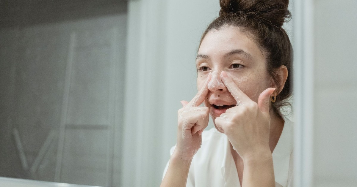 A young woman applies skincare in front of a bathroom mirror, focusing on facial care.