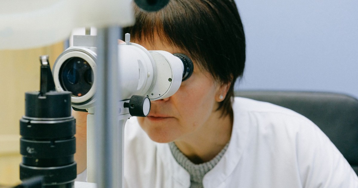 Anonymous female biologist in white coat observing object through big lab microscope while sitting on grey chair and leaning forward in light workplace