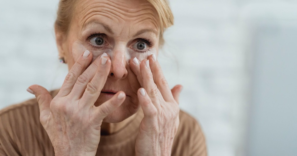 Crop focused mature female applying moisturizing nourishing eye patches and looking away in light bathroom